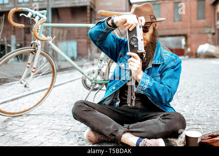 Tanga barbuto vestito elegantemente con il cappello e giacca seduta con foto e fotocamera retrò bicicletta su strada all'aperto Foto Stock