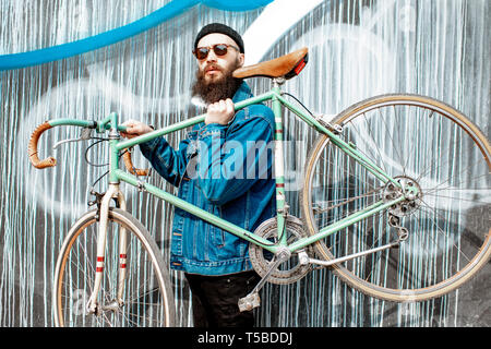 Ritratto di un elegante uomo barbuto vestito in giacca e cappello con bicicletta retrò sulla parete colorata sullo sfondo Foto Stock