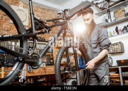 Bello barbuto repairman in abbigliamento da lavoro di montaggio sulla ruota di una bicicletta di montagna in officina Foto Stock