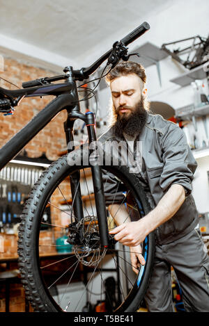 Bello barbuto repairman in abbigliamento da lavoro di montaggio sulla ruota di una bicicletta di montagna in officina Foto Stock