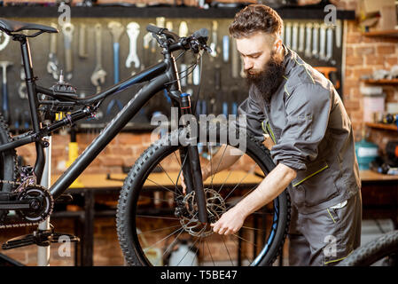 Bello barbuto repairman in abbigliamento da lavoro di montaggio sulla ruota di una bicicletta di montagna in officina Foto Stock