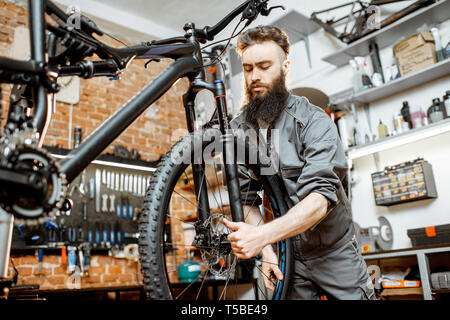 Bello barbuto repairman in abbigliamento da lavoro di montaggio sulla ruota di una bicicletta di montagna in officina Foto Stock