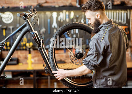 Bello repairman in workwear serve bicicletta di montagna, in piedi con la ruota anteriore in officina di un negozio di biciclette Foto Stock