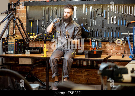 Ritratto di un bel repairman in workwear seduta con chiavi dinamometriche e diversi strumenti di lavoro in officina di biciclette Foto Stock