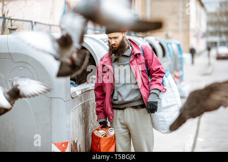 Ritratto di un senzatetto premuto mendicante in piedi con i sacchetti vicino al cestino di contenitori in città. Concetto di povertà e disoccupazione Foto Stock