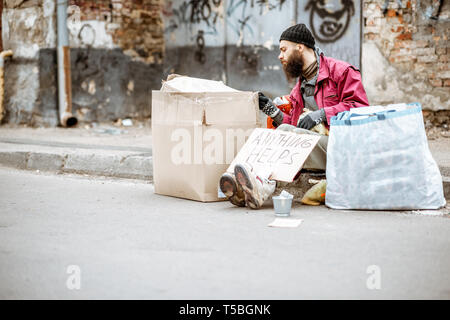 Senzatetto premuto mendicante seduto con i sacchetti e cartoni sulla strada sulla parete frastagliata sfondo. Concetto di povertà sociale Foto Stock