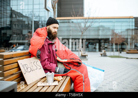 Senzatetto e disoccupati mendicante seduto sul banco avvolto con sacco a pelo di accattonaggio denaro vicino al centro business Foto Stock