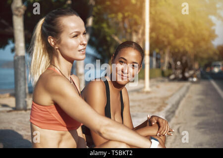 Sorridente giovane donna in sportswear seduti su un marciapiede con il suo partner di allenamento prendendo una pausa dalla loro esecuzione Foto Stock