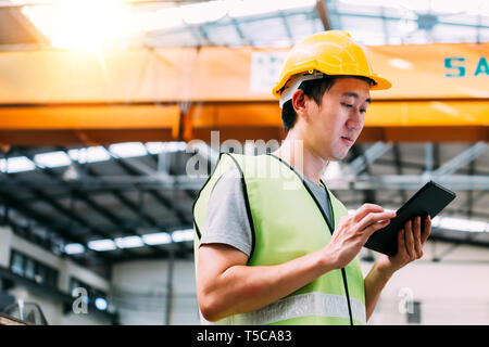 Giovani asiatici pesante maschio industral lavoratore utilizzando una tavoletta digitale all'interno della fabbricazione e del sito di fabbrica con gru dietro Foto Stock