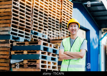 Sorridente e felice asiatici industriale magazzino adulto lavoratore dotato di compressori hardhat e uniforme di posti con le braccia incrociate nella parte anteriore della distribuzione logistica Foto Stock