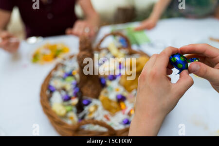 Le mani di un bambino scartare una caramella di cioccolato uovo con un grande uovo di pasqua cesto in background durante le festività di Pasqua cellebrations con familiari e parenti Foto Stock