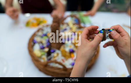 Le mani di un bambino scartare una caramella di cioccolato uovo con un grande uovo di pasqua cesto in background durante le festività di Pasqua cellebrations con familiari e parenti Foto Stock