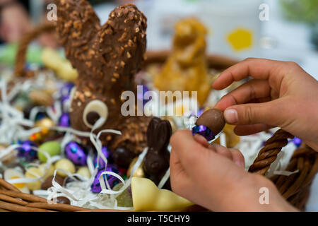 Le mani di un bambino scartare una caramella di cioccolato uovo con un grande uovo di pasqua cesto in background durante le festività di Pasqua cellebrations con familiari e parenti Foto Stock