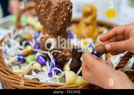 Le mani di un bambino scartare una caramella di cioccolato uovo con un grande uovo di pasqua cesto in background durante le festività di Pasqua cellebrations con familiari e parenti Foto Stock