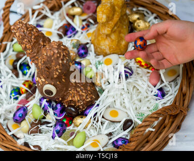 Le mani di un bambino scartare una caramella di cioccolato uovo con un grande uovo di pasqua cesto in background durante le festività di Pasqua cellebrations con familiari e parenti Foto Stock