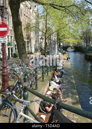 Vista di una scena del canale nel centro di Amsterdam con gli alti edifici lungo il canal, lotti di biciclette parcheggiate e persone godendo il sole di aprile Foto Stock