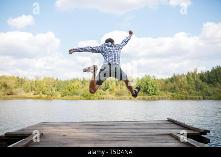 Foto dal retro del giovane uomo salto sul ponte in legno sul fiume Foto Stock