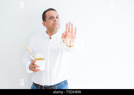 La mezza età uomo mangiare spaghetti asiatici da portar via cartone con mano aperta facendo segno di stop con gravi e fiducioso espressione, gesto di difesa Foto Stock