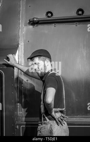 Bianco e nero close up della locomotiva a vapore cabina equipaggio. Driver del treno nel tappo piatto & tute, in piedi sulla piattaforma appoggiata contro il lato del motore a vapore. Foto Stock