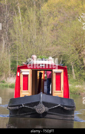 Ritratto di narrowboat che sopraggiungono sul Regno Unito canal, un pensionato coppia sposata in corrispondenza della parte posteriore della barca, rivolto in avanti, sterzo rosso brillante canal boat in sun. Foto Stock