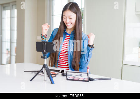 Bella asiatica influencer donna la registrazione compongono tutorial utilizzando la fotocamera dello smartphone molto felici ed eccitati facendo vincitore gesto con le braccia sollevate, SMIL Foto Stock