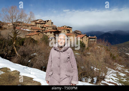 Una donna anziana gode di un paesaggio in un altopiano autentico villaggio catalano con case in pietra mentre si cammina lungo un sentiero di montagna ricoperta di neve di fusione. Foto Stock