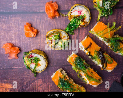 Vista dall'alto di piatti giapponesi su sfondo scuro. Sushi di salmone, sushi rotoli a bordo, carne di granchio, gamberetti, wakame, lo zenzero, la superficie in legno Foto Stock