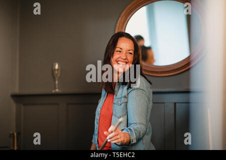 Ritratto di un sorridente metà donna adulta, sta in piedi in ambienti chiusi tenendo un pool cue e ridere. Foto Stock
