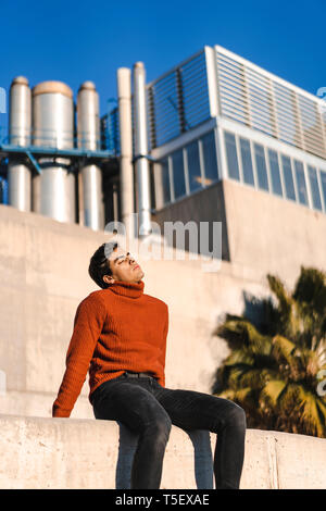 Ritratto di moda giovane uomo che indossa turtleneck pullover seduto su una parete a luce solare rilassante Foto Stock