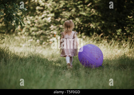 Eleganti bambina 2-5 anni azienda grande mongolfiera indossando trendy abito rosa in prato. Giocoso. Festa di compleanno. bambina con un palloncino in par Foto Stock