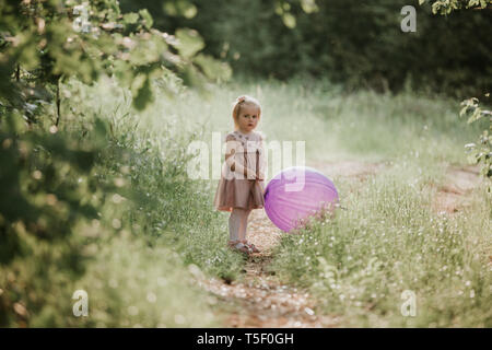 Eleganti bambina 2-5 anni azienda grande mongolfiera indossando trendy abito rosa in prato. Giocoso. Festa di compleanno. bambina con un palloncino in par Foto Stock