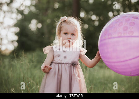 Eleganti bambina 2-5 anni azienda grande mongolfiera indossando trendy abito rosa in prato. Giocoso. Festa di compleanno. bambina con un palloncino in par Foto Stock