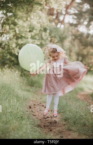 Eleganti bambina 4-5 anno vecchia azienda grande mongolfiera indossando trendy abito rosa in prato. Giocoso. Festa di compleanno. bambina con un palloncino in par Foto Stock