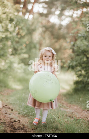 Eleganti bambina 4-5 anno vecchia azienda grande mongolfiera indossando trendy abito rosa in prato. Giocoso. Festa di compleanno. bambina con un palloncino in par Foto Stock