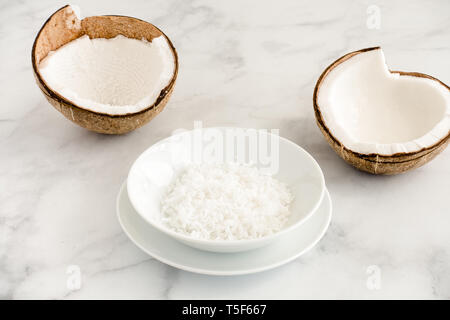 Due metà di noce di cocco e scaglie di noce di cocco in un colore bianco porcellana ciotola su marmo bianco sfondo con copia spazio. Angolo di visualizzazione. Foto Stock