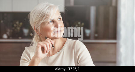 Allegro senior ritratto di donna. Titolare di pensione o di rendita donna in possesso di una penna, si siede a casa e guarda la finestra. Concetto di un felice pensionamento Foto Stock