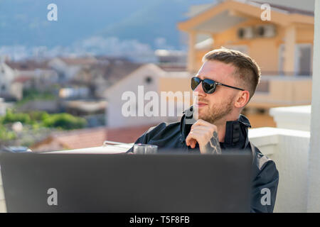 Bello pensieroso elegante uomo seduto sul balcone e di lavoro sul computer portatile. Home office. Concetto di freelance. Giovane imprenditore. Foto Stock