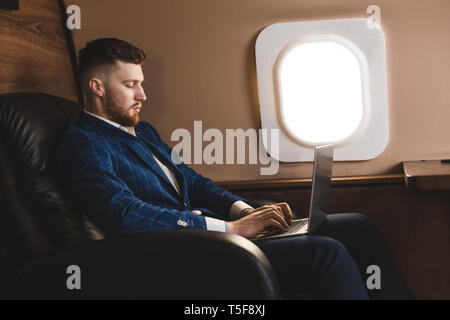 Attraente e imprenditore di successo lavorando su un computer portatile mentre è seduto in una sedia del suo jet privato. Foto Stock