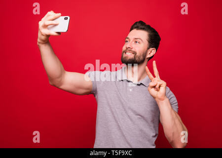 Giocoso uomo barbuto rendendo selfie sullo smartphone e mostrando il pollice in alto su sfondo rosso Foto Stock