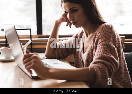 Attraente giovane donna siede a un tavolo in una caffetteria con una tazza di caffè e gode di un laptop Foto Stock