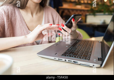 Attraente giovane donna siede a un tavolo in una caffetteria con una tazza di caffè e un laptop e usa il telefono Foto Stock