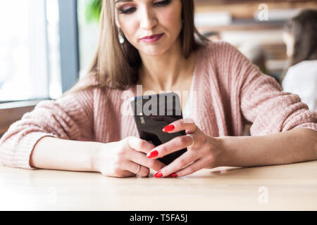 Bella giovane donna si siede a un tavolo in un bar con una tazza di caffè e utilizza il telefono Foto Stock