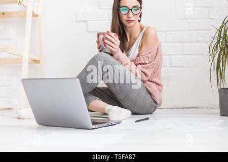 Una giovane donna felice si siede sul pavimento in un luminoso appartamento o di un ufficio interno e lavora su un portatile, freelancer ragazza al lavoro, il concetto di distanza Foto Stock