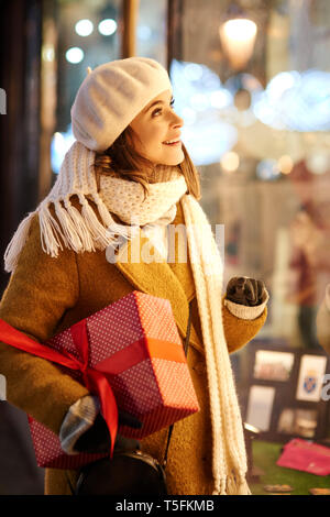 Donna sorridente con il regalo di Natale cercando nella finestra del negozio Foto Stock