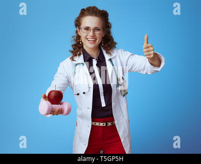 Felice medico moderno donna in bue t-shirt, pantaloni rosso e bianco manto medica con il manubrio e apple che mostra i pollici in su contro sfondo blu. Foto Stock