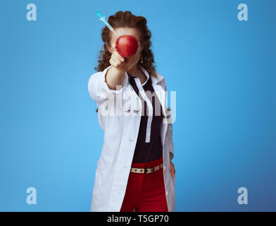 Elegante medico donna in bue t-shirt, pantaloni rosso e bianco manto medico tenendo un Apple con una siringa bloccato nella parte anteriore della faccia isolato Foto Stock