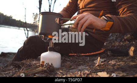 Turismo a tema escursioni e viaggi nella natura. Mani uomo caucasico utilizza attrezzando per cuocere i cibi fuori. luoghi turistici di pentola di acqua sul cilindro di bruciatore Foto Stock