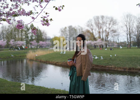 Giovane donna di bere il caffè da un bicchiere di carta che indossa color smeraldo mantello - sakura colorati fiori di ciliegio in un parco a Riga, a est della capitale europea Foto Stock