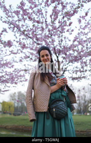 Giovane donna di bere il caffè da un bicchiere di carta che indossa color smeraldo mantello - sakura colorati fiori di ciliegio in un parco a Riga, a est della capitale europea Foto Stock