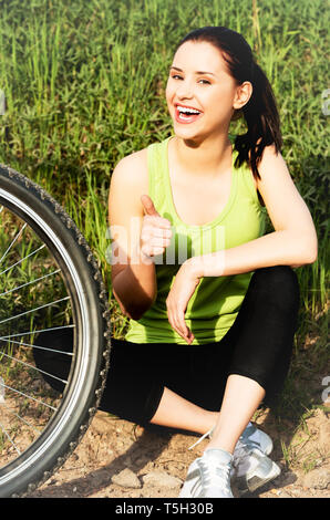 Giovane donna appoggiata sul terreno dopo la bicicletta equitazione e rendendo tumbs up. Foto Stock